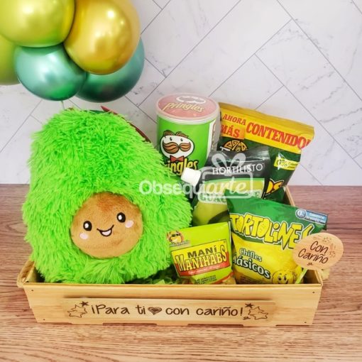 Una caja de regalo llena de guacamole con bocadillos, globos y un peluche de aguacate verde.