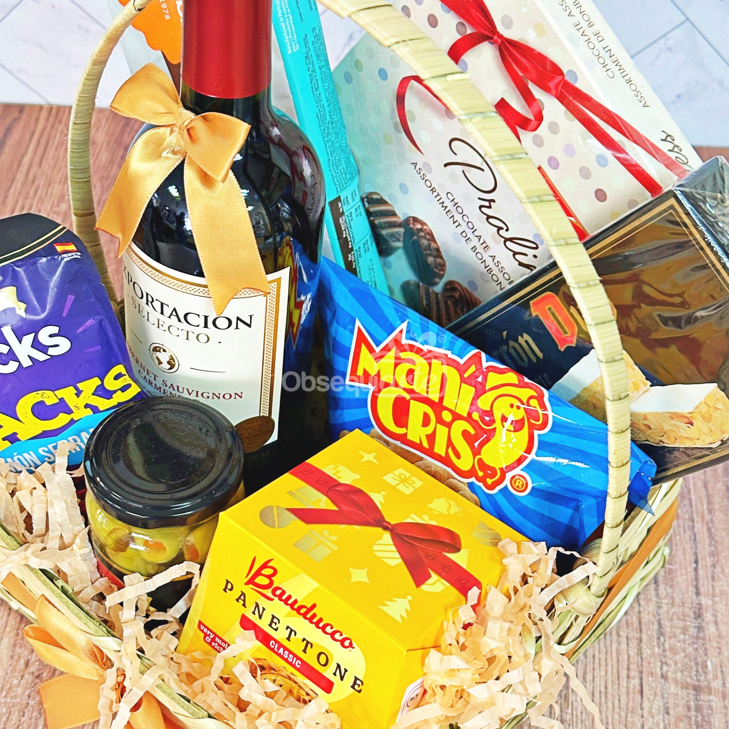El Regalo Canasta Festiva incluye una botella de vino tinto, bombones, galletas, panettone en caja amarilla, aperitivos y aceitunas, todo ello elegantemente presentado en la cesta de regalo.