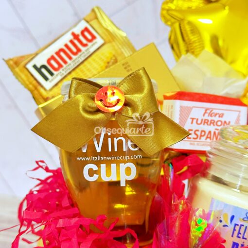 El paquete de regalo Regalo Primavera con Vela y Vino incluye una copa de vino adornada con un lazo dorado, una oblea Hanuta, un turrón español Flora y una vela, todo bellamente arreglado con papel decorativo colorido para un toque elegante.
