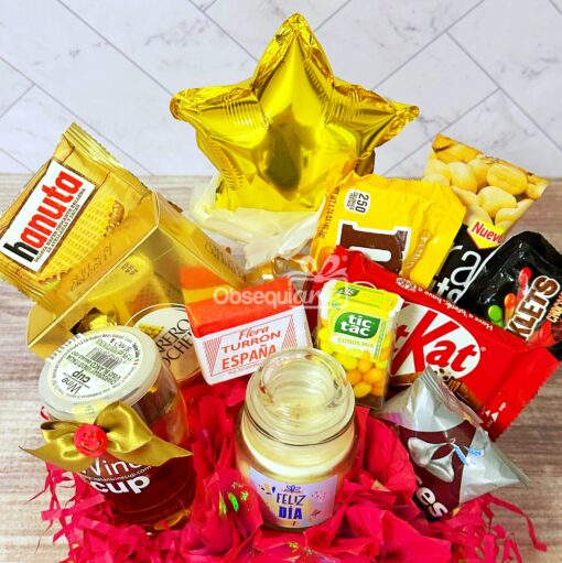 Una cesta de regalo de Regalo Primavera con Vela y Vino repleta de deliciosos snacks como Hanuta, KitKat y M&M's. Incluye un tarro, un globo con forma de estrella y una botella pequeña, todos bellamente dispuestos en un encantador papel rosa para capturar la esencia de la alegría primaveral.