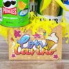La canasta Regalo Fiesta incluye una lata de Pringles, una cerveza y una tarjeta decorativa con "Con cariño" e ilustraciones vibrantes. La cesta está llena de papel decorativo amarillo.