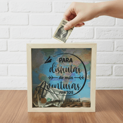 Una mano introduce un billete de un dólar en la caja de sombra Regalo Alcancia Vamos Juntos, que tiene dinero ahorrado, un fondo de mapamundi y un texto en español sobre ahorrar para vivir aventuras juntos.