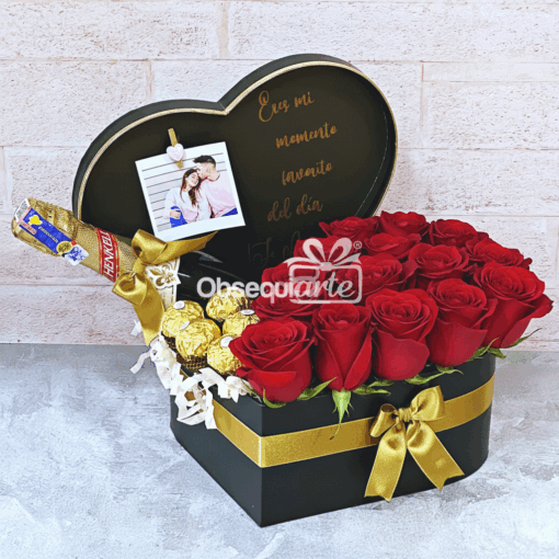 Regalo Corazón de Rosas: Una caja negra en forma de corazón con un lazo dorado, llena de rosas rojas, bombones Ferrero Rocher, un KitKat y una foto de pareja, sobre un fondo de ladrillo claro.