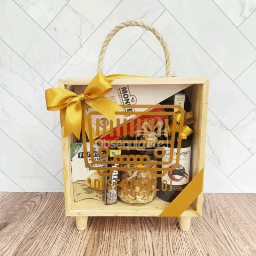 La caja de regalo Regalo Brunch Vintage cuenta con un asa de cuerda, un lazo amarillo y un surtido de aperitivos y bebidas en su interior, presentados sobre una superficie de madera con fondo de azulejos.
