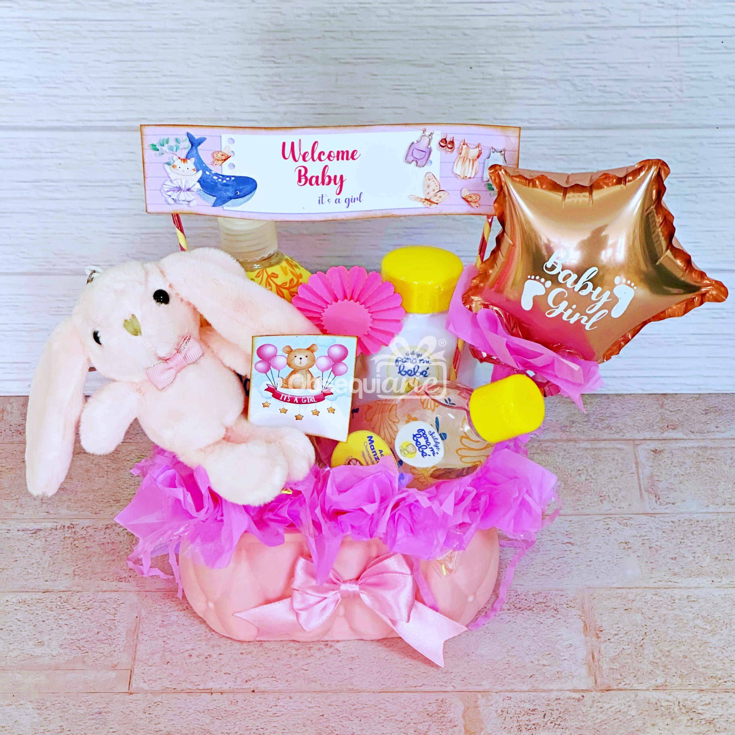 La cesta de regalo Regalo Bienvenida Bebé (Niña) incluye un conejito de peluche, artículos para el cuidado del bebé, un cartel de "Bienvenida Bebé", un globo de "Niña" y lazos rosas, perfectos para celebrar la llegada de un nuevo bebé.