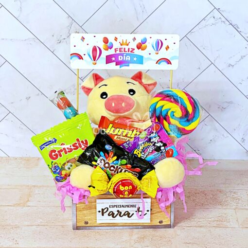 Una caja de madera que contiene un cerdo de peluche, paquetes de dulces, una paleta colorida y un cartel de "Feliz Día", lleno de papel triturado rosa: el Regalo Abrazo Dulcero perfecto para cualquier celebración.