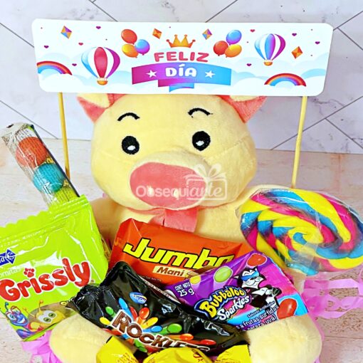 Un juguete de peluche con un cartel que dice "Feliz Día" está rodeado de una variedad de dulces, incluidos Grissly, Jumbo, Rocklets y Bubbaloo, y una paleta grande. En el cartel se representan globos de colores. Este dulce Regalo Abrazo Dulcero es perfecto para cualquier ocasión.