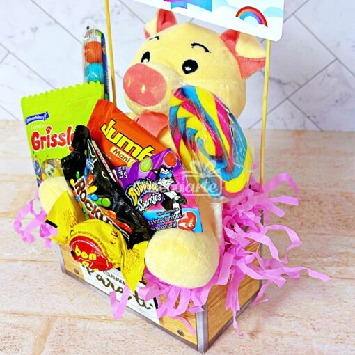 Un delicioso Regalo Abrazo Dulcero que contiene un cerdito de peluche para un abrazo acogedor, varios dulces, una piruleta y un relleno de papel triturado rosa. La canasta se asienta sobre una superficie de color claro con un fondo de azulejos geométricos, lo que la hace perfecta para cualquier ocasión dulce.