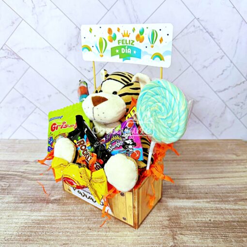 Un Regalo Abrazo Dulcero Rosado (copia) que incluye un tigre de peluche, dulces variados, una piruleta grande y un cartel que dice "FELIZ DÍA", todo dispuesto en una caja de madera con papel naranja triturado.