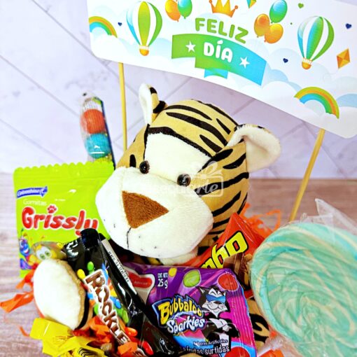 Un delicioso Regalo Abrazo Dulcero Rosado (copia) que presenta un tigre de peluche, dulces variados, una piruleta en espiral verde y un letrero que dice "Feliz Día" en español, todo decorado con globos y nubes.