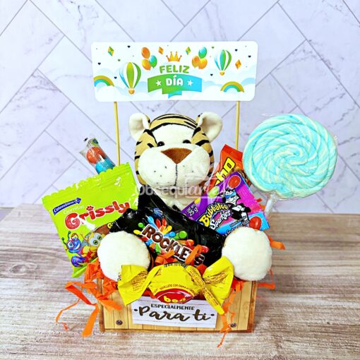 Canasta de madera con un pequeño tigre de peluche, varios paquetes de dulces, una piruleta grande y un cartel de "Feliz Día" decorado con globos y arcoíris en el fondo: el Regalo Abrazo Dulcero Verde perfecto