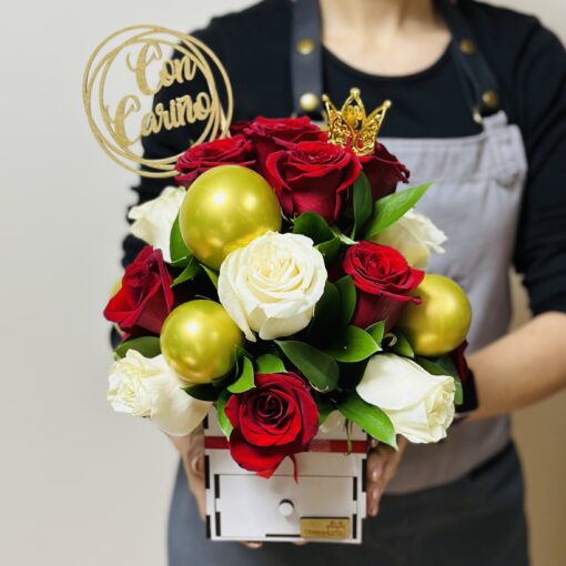 Una persona con un delantal oscuro sosteniendo un ramo de rosas rojas y blancas, adornado con bolas doradas y una mariposa, con un cartel de "Regalo Soft Queen".