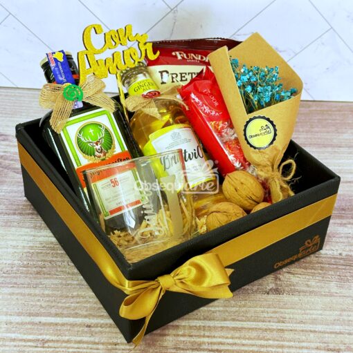Una caja Regalo Élite que incluye una botella de licor, un vaso, nueces, bocadillos, un pequeño ramo y un cartel decorativo "Con Amor", todo elegantemente envuelto con una cinta dorada.