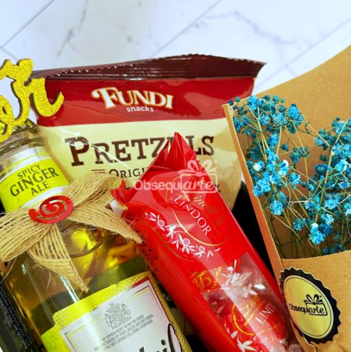 Descubra la caja Regalo Élite: un arreglo de regalo seleccionado que presenta ginger ale picante, pretzels, chocolates Lindor y flores de color azul intenso, colocados con buen gusto sobre una superficie de mármol.