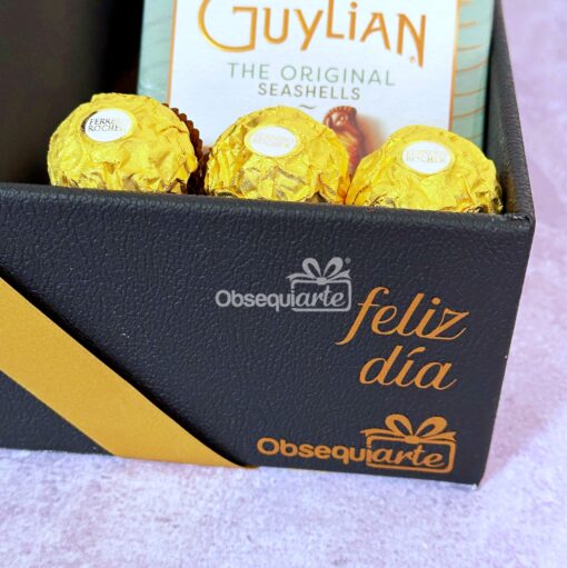 Caja de regalo negra con lazo dorado que contiene bombones Ferrero Rocher y una caja de conchas Guylian. El texto de la caja dice "feliz día" y "Obsequiarte".