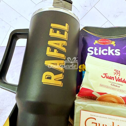 Una taza de viaje negra con la palabra "RAFAEL" escrita en ella, junto a una bolsa de Mallorca Sticks, un paquete de café Juan Valdez y un té de ginseng.