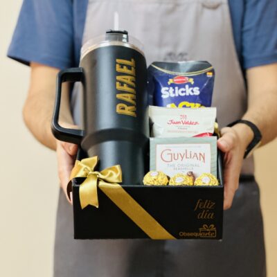 Una persona sostiene una caja de regalo de Regalo Prestige, que muestra un vaso negro personalizado, una variedad de bocadillos y está adornada con un lazo dorado.