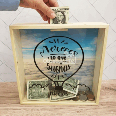 Una mano introduce un dólar doblado en el Regalo Alcancía Cumple Sueños, una caja de madera con monedas y billetes en su interior. La caja muestra un globo aerostático y la frase: 