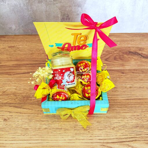 El Regalo Luminé con Vela incluye una tarjeta "Te Amo", un frasco de dulces, chocolates envueltos en amarillo atados con cintas rojas y amarillas y una vela Luminé sobre una superficie de madera.