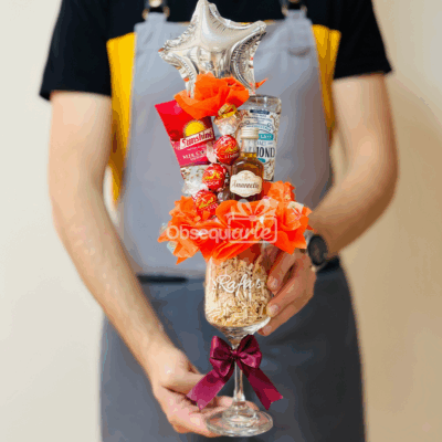 Una persona vestida con delantal sostiene un Regalo Copa Elegante, una gran copa de vino que contiene bombones, una botella de licor en miniatura, papel picado de relleno, un adorno y está adornada con un lazo decorativo.