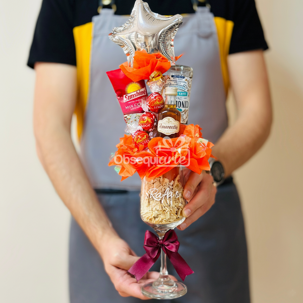 Una persona vestida con delantal sostiene un Regalo Copa Elegante, una gran copa de vino que contiene bombones, una botella de licor en miniatura, papel picado de relleno, un adorno y está adornada con un lazo decorativo.