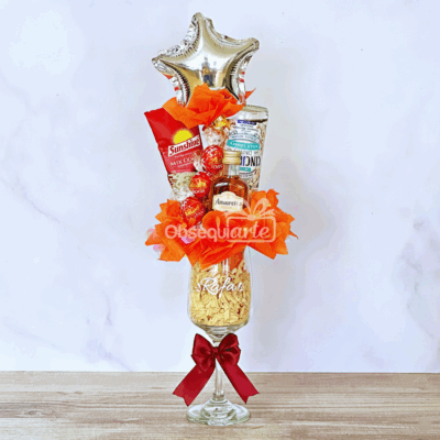 El vaso alto Regalo Copa Elegante, decorado con un lazo rojo y un envoltorio naranja, contiene bombones variados, una bebida en lata y un globo plateado en forma de estrella.