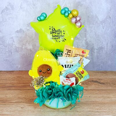 Una cesta de regalo con aperitivos, un Regalo Taza Sopera, un globo amarillo con forma de estrella, un aguacate de peluche y papel de seda verde azulado, todo ello sobre un fondo gris claro.