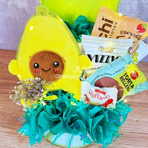 Una encantadora cesta de regalo con un aguacate de peluche, aperitivos, caramelos Raffaello, una barrita de cereales, flores decorativas y el delicioso Regalo Taza Sopera, bellamente dispuestos en papel de seda verde.