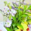 El "Ramo de Flores Brisa de Alegría" presenta una vibrante variedad de flores de color violeta claro, amarillo y violeta oscuro con exuberantes hojas verdes, elegantemente envueltas en papel blanco, perfectas para agregar alegría a cualquier ocasión.