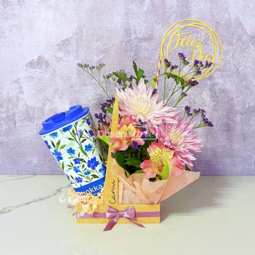 La taza "Regalo Mug con Flores" presenta un colorido arreglo floral de flores rosas y moradas en una caja "Connie", combinada con una taza azul con estampado floral y un adorno "Feliz Día", creando el regalo perfecto para alegrar el día de cualquier persona.
