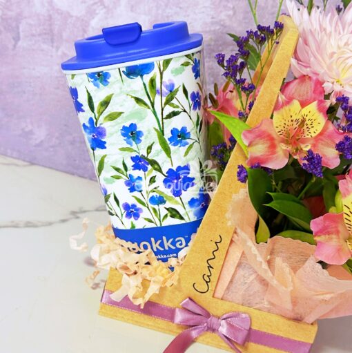 La taza Regalo con Flores, con diseño floral y tapa azul, combina perfectamente con un ramo de flores rosas y amarillas envueltas en papel decorativo, creando un regalo ideal.
