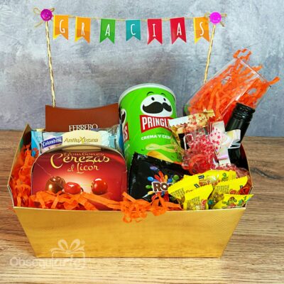 La cesta Regalo Full Feliz está llena de aperitivos, caramelos, una lata de Pringles y una botellita. Está decorada con papel naranja y rematada con una pancarta de 