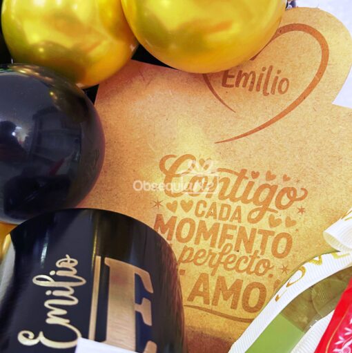 Regalo Galaxy (copia): Una tarjeta de madera grabada con un corazón, el nombre "Emilio" y texto en español, combinada con globos negros y dorados y un vaso negro con una "E" dorada, crea un elegante conjunto de regalo.