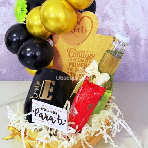 Regalo Galaxy (copia): Set de regalo con globos negros y dorados, una taza con la etiqueta "Emilio", un elegante vaso, dos tubos de loción y una placa de madera grabada dispuesta en una bandeja de madera.