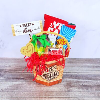 La canasta de regalo incluye bocadillos variados, una cinta verde, una etiqueta con forma de corazón, papel triturado rojo y un cartel de madera que dice 