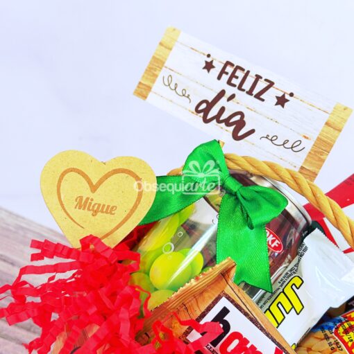 Canasta de regalo con bocadillos, un lazo verde, relleno de papel rojo, un corazón de madera con la etiqueta "Migue" y un cartel en la parte posterior que dice "Feliz día".