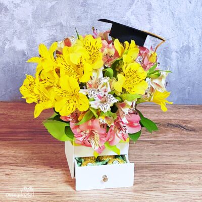 Regalo Soft con Astromelias: Un arreglo compacto de astromelias amarillas y rosas con un birrete de graduación, presentado en una caja blanca con un cajón abierto lleno de bombones, perfecto para celebrar los logros.