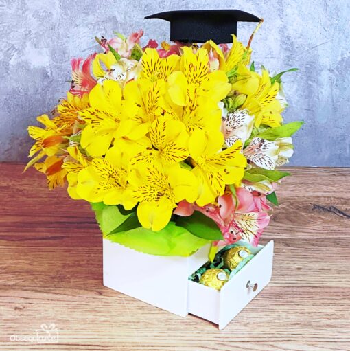 El Regalo Soft con Astromelias presenta Astromelias amarillas y rosas en una caja blanca con un gorro de graduación en la parte superior, además de un cajón lleno de deliciosos bombones.