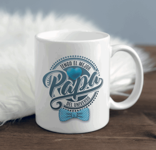 Taza blanca con gráfico de corazón y moño azul, texto en español "Tengo el mejor papá del universo" (el Regalo Taza Super Papá (copia), que se muestra sobre una superficie de madera con un fondo blanco suave.