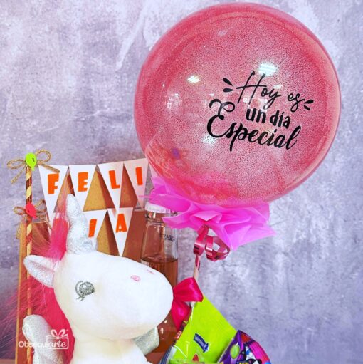 El "Regalo Bandeja Feliz Día con Unicornio" incluye un globo rosa con texto en español, un unicornio de peluche, aperitivos y banderas triangulares con la inscripción "FELICIDAD", todo ello bellamente dispuesto sobre un fondo gris.