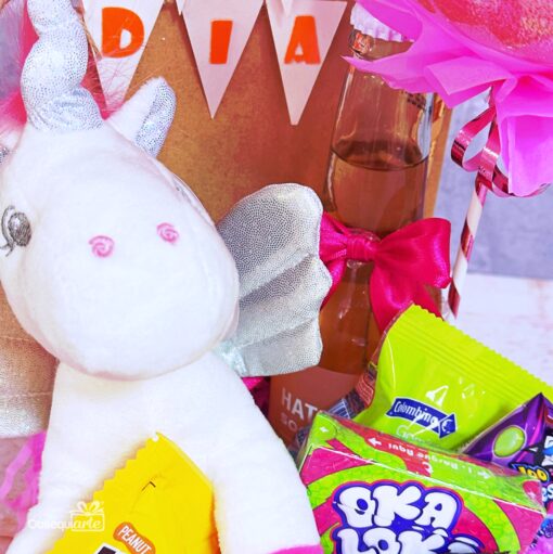 El Regalo Bandeja Feliz Día con Unicornio incluye un peluche de unicornio blanco, caramelos de cacahuete, caramelos Oka Loka verdes y azules, una botella con un lazo rosa y decoración de fiesta con una pancarta "Bandeja Feliz Día", ideal para cualquier celebración.