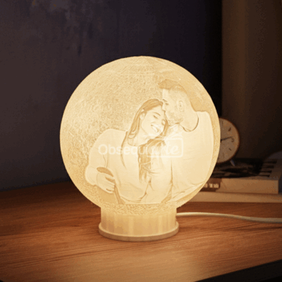 La Regalo Lampara 3D Moon Personalizada, con la imagen grabada de una pareja sonriente, descansa sobre madera junto a un reloj y libros, siendo el detalle ideal para un regalo único y especial.