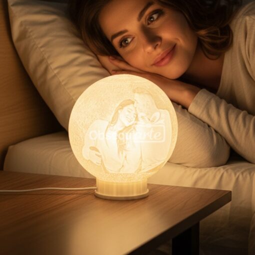 Una mujer en la cama sonríe ante una brillante Lámpara Regalo 3D Moon Personalizada en su mesilla de noche, con una imagen grabada a medida de una pareja feliz, perfecta para hacer que los momentos especiales sean inolvidables.