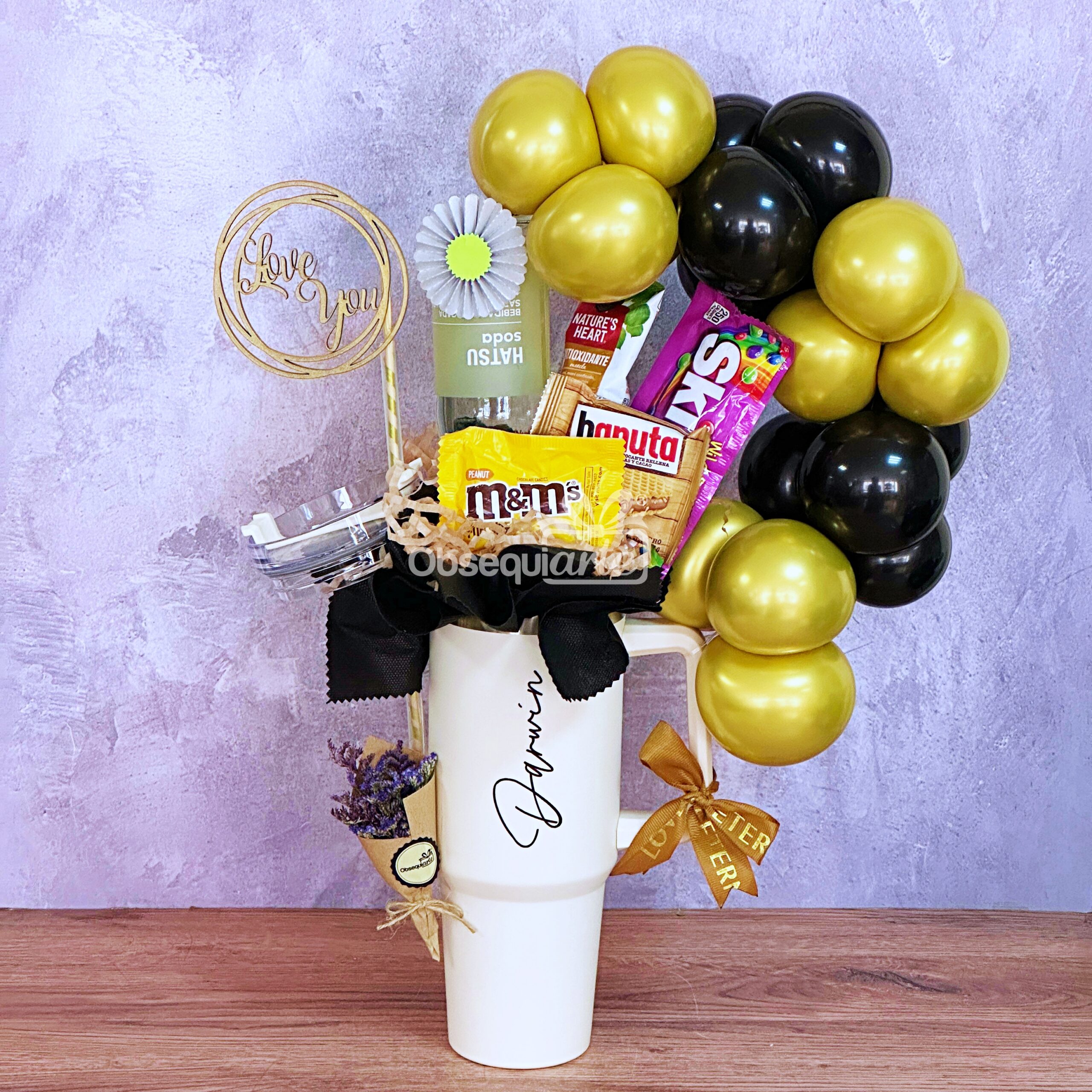 Regalo Essence para Hombre: Incluye una taza blanca con dulces surtidos, esencia chave de sabor, pequeño ramo y globos dorados y negros, presentado sobre fondo púrpura. Ideal como regalo para hombre.