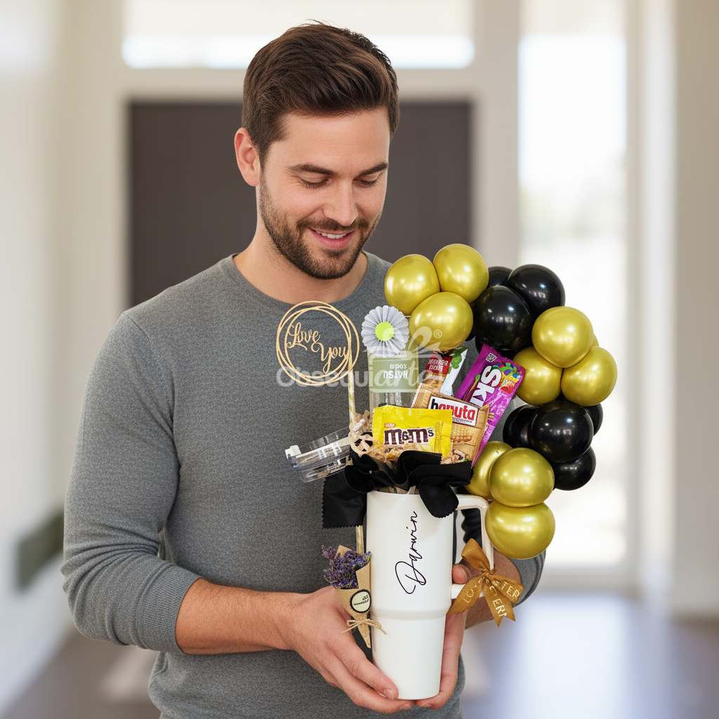 Un hombre sostiene el "Regalo Essence para Hombre" en el interior, con una taza blanca de regalo llena de caramelos variados, globos dorados y negros, y una decoración festiva que capta el espíritu de un regalo atento.