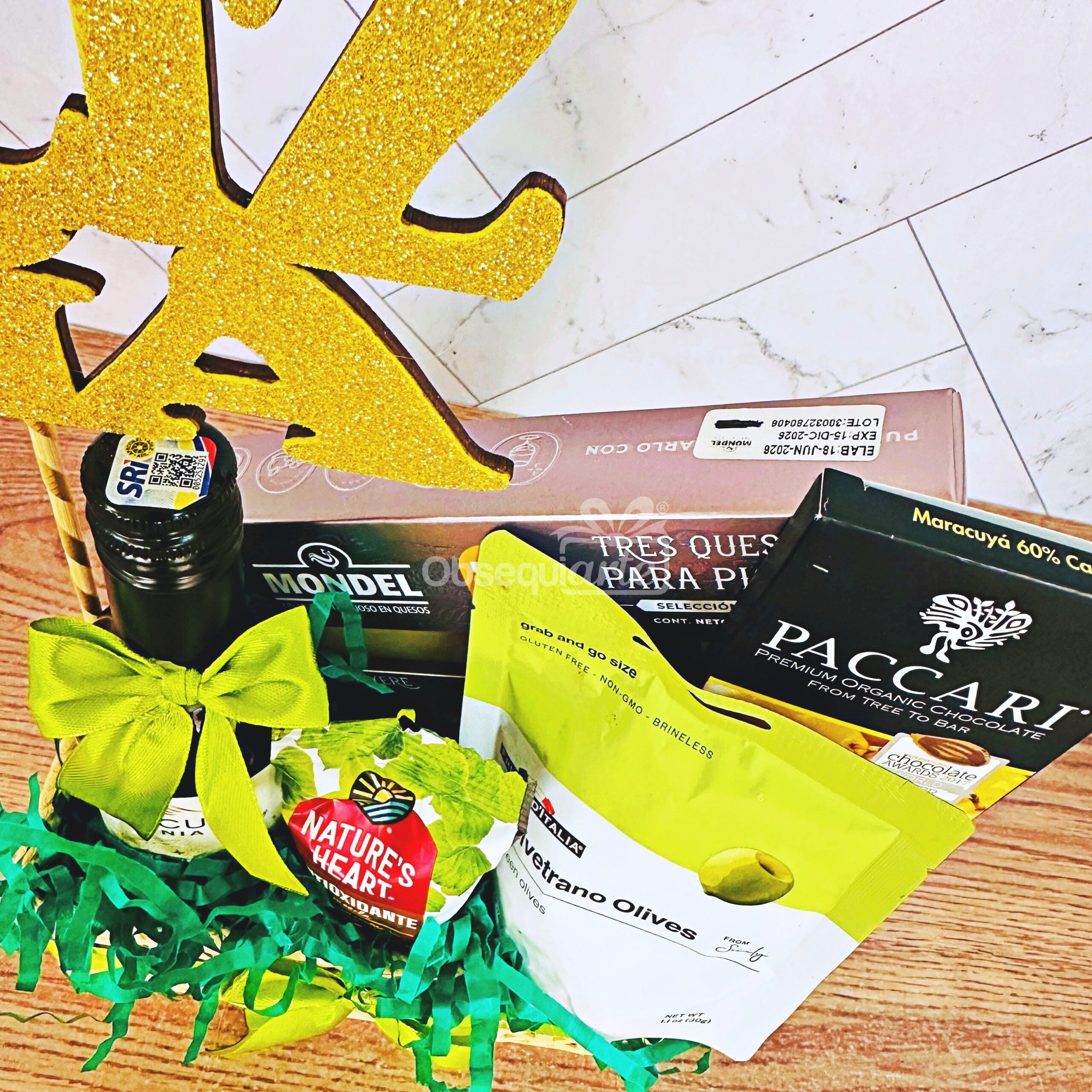 La cesta de regalo «Regalo Para Disfrutar» incluye chocolate, queso, aceitunas, aceite de oliva, un paquete de aperitivos y un adorno dorado brillante, todo ello bellamente dispuesto sobre una base de madera clara.