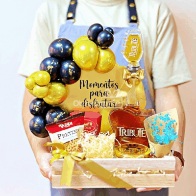 Una persona sostiene una cesta Regalo Royal Guitar con whisky Tribute, aperitivos, una manzana de caramelo azul, globos dorados y negros y un cartel que dice 