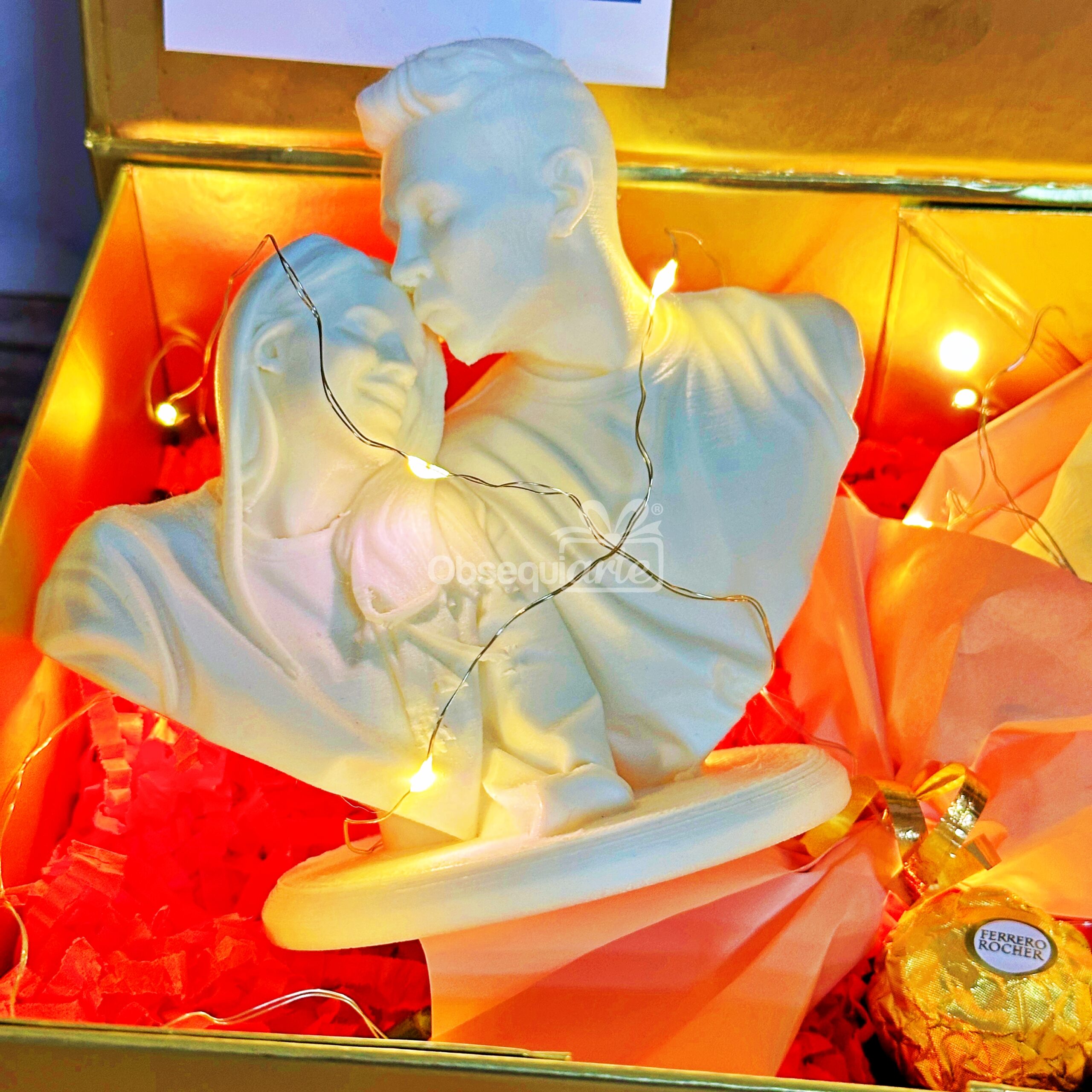 La escultura Regalo Amor Eterno de busto blanco de una pareja está bellamente presentada en una caja de regalo con luces de hadas, papel de seda naranja y chocolate Ferrero Rocher.