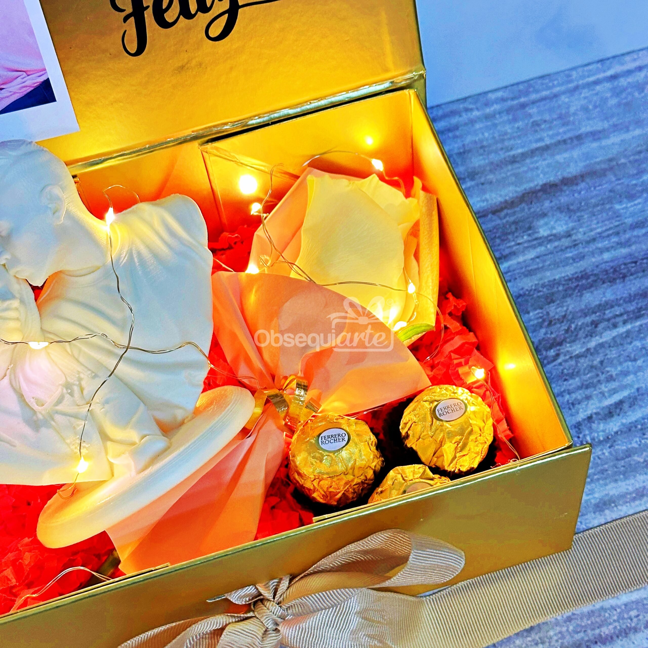 Regalo Amor Eterno: Una caja dorada con una estatua blanca, una rosa amarilla que simboliza el amor eterno, bombones Ferrero Rocher, luces de cuerda, relleno de papel rojo y un lazo beige.