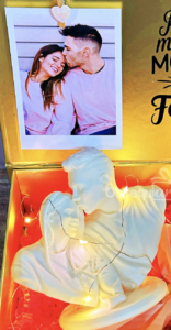 Una foto enmarcada de una pareja sonriente aparece sobre una escultura blanca de una pareja y un niño, rodeada de pequeñas luces de cuerda y papel decorativo rojo.